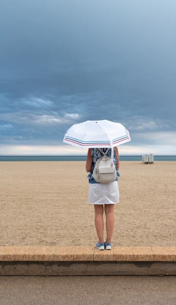 A photo of a person carrying folded beach chairs and umbrella on a sunny beach, smiling, and walking towards the sea.