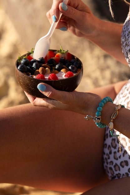 Close-up of a person's hand holding a colorful fruit skewer with strawberries, blueberries, and kiwi, with the beach and ocean blurred in the background. The sun is shining brightly.