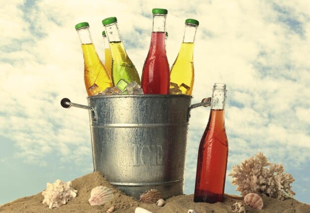 A picturesque beach scene with a cooler filled with assorted canned and bottled beverages, highlighting a relaxed and convenient beach outing. The view includes beach chairs, towels, and a sun umbrella.