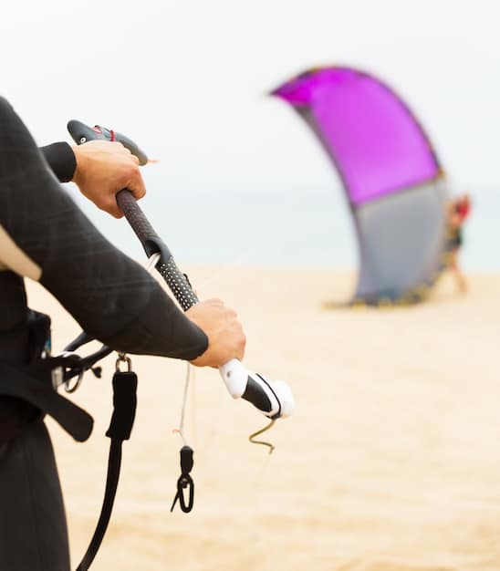 A close-up of a kitesurfer's quick-release system on their harness, showing the mechanism and how it's engaged. The image is well-lit and clearly shows the different parts of the system.