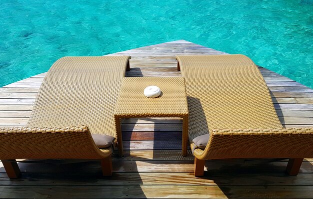 A close-up shot of the deck of an overwater bungalow, showcasing luxurious outdoor furniture, including a plush daybed and a table set for two. The turquoise water is visible below, and the scene exudes tranquility and romance.