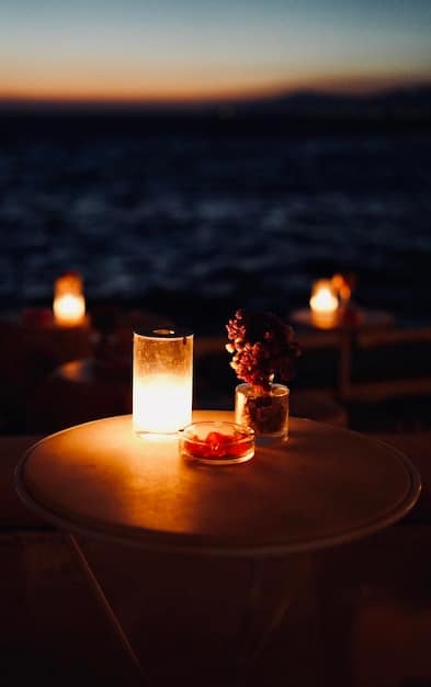 An elegant cocktail being served on a luxury beach resort VIP lounge table. The background features a soft-focus view of the beach and ocean at sunset.