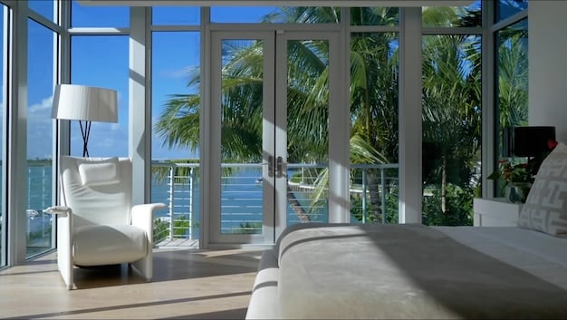 A well-appointed interior of a luxury beach resort suite. Large glass doors open onto a balcony with an ocean view, but sheer curtains filter the sunlight, creating a soft, diffused glow. Light colored walls and furnishings enhance the bright yet cool ambiance of the room.