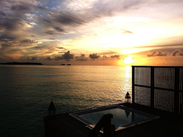 A scenic view from a high-floor suite at a luxury beach resort at sunset. The sky is painted with vibrant oranges, pinks, and purples. Sunlight reflects off the ocean surface, creating a dazzling effect. The balcony is partially shaded, providing a comfortable viewing spot.
