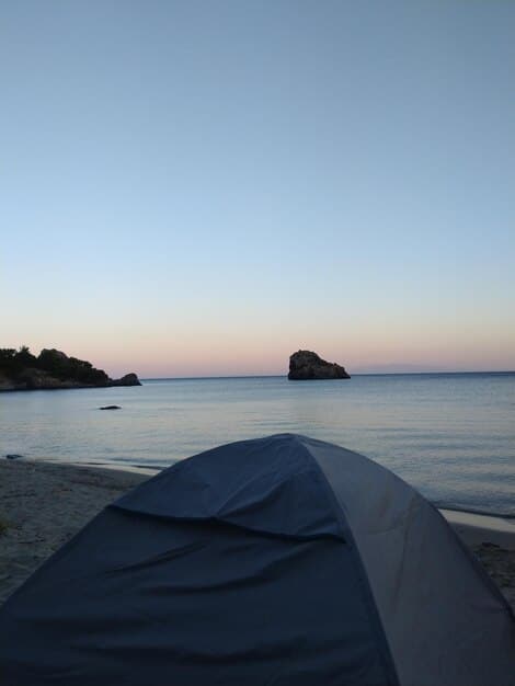 A serene beach campsite at dusk, showing a simple tent set up on the sand, with the ocean waves gently lapping the shore in the background.  Focus should be on the peaceful setting and natural beauty.
