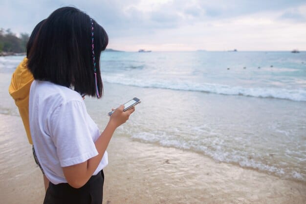 A person standing on a beach, looking out at the ocean with a smartphone in hand, possibly checking travel deals, booking flights, or managing loyalty points.