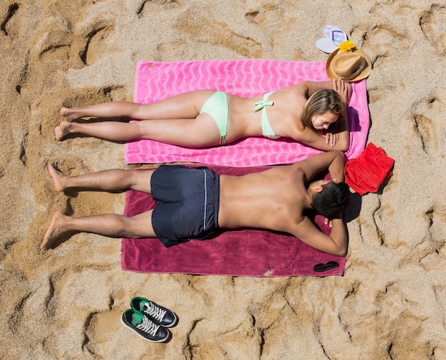 A vibrant scene of beachgoers enjoying sunbathing and swimming. Some people are relaxing on towels with umbrellas providing shade, while others are splashing and playing in the clear, shallow water. Beach toys and colorful accessories are scattered around, enhancing the cheerful atmosphere.