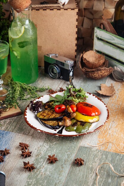 A close-up shot of fresh, colorful seafood and vegetables laid out on a wooden picnic table, ready to be grilled. The backdrop includes a blurry beach scene at sunset.