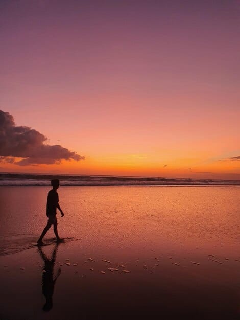 A vibrant sunset over a calm beach, with a single silhouette of a person walking along the shoreline. Various pastel colors paint the sky, reflecting beautifully on the wet sand. A few scattered seashells are visible close to the viewer.