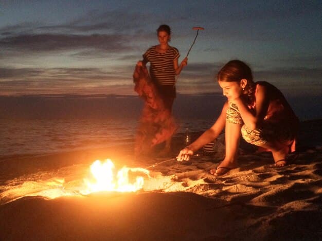 A lively scene at a beach bonfire at twilight, with people gathered around, laughing and talking. Soft, warm light illuminates the scene. In the background, the ocean waves gently roll in, and a few stars start to appear in the darkening sky.