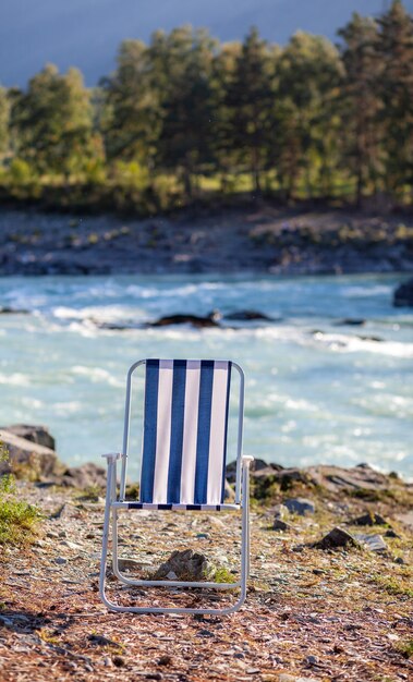 A close-up of a foldable beach chair with a drink holder, positioned on a sandy beach with the ocean in the blurred background. The chair is lightweight and portable.