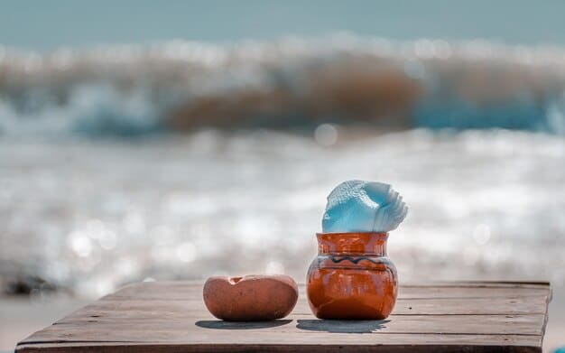 A close-up shot of a reusable water bottle and a small cooler filled with ice packs and various drinks on a beach towel. Sand and seashells are scattered nearby.