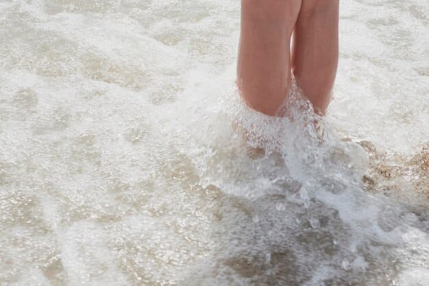 A close-up shot of a person's bare feet sinking into soft, wet sand at the water's edge, with gentle waves approaching, evoking a sense of calm and sensory connection to the beach.