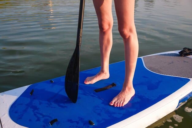 A close-up of a paddleboarder's bare feet standing on the textured deck pad of a paddleboard, with calm blue water visible around the edges of the board. The sun is reflecting gently off the water.