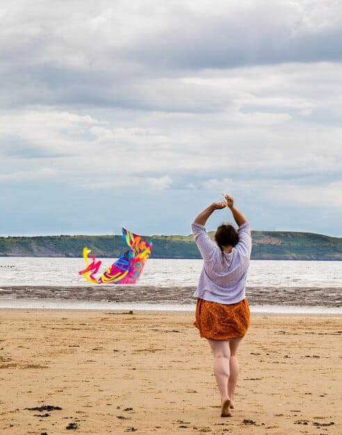 A person happily flying a colorful kite on a wide, sandy beach with the ocean in the background, illustrating active and joyful recreation during a beach camping trip.