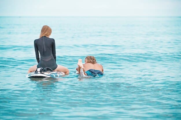 A child joyfully bodyboarding on a small, gentle wave, with an adult supervising closely in the shallow water, clear blue sky and distant shoreline in the background.