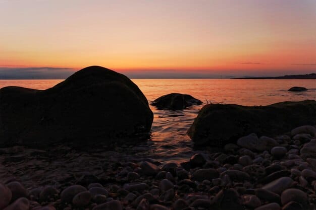 A secluded, rocky cove beach at sunset, with golden light reflecting on the calm water and dark silhouettes of rock formations. The sky displays soft hues of orange and purple.