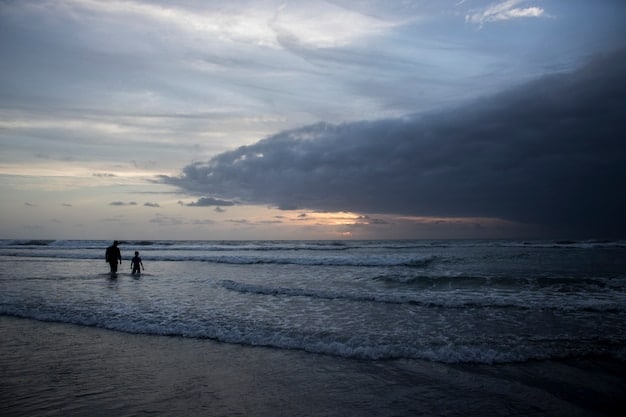 A dark, moody sky over an ocean with large waves beginning to form, indicating a sudden change in weather, with a lone person quickly packing their beach gear in the foreground.