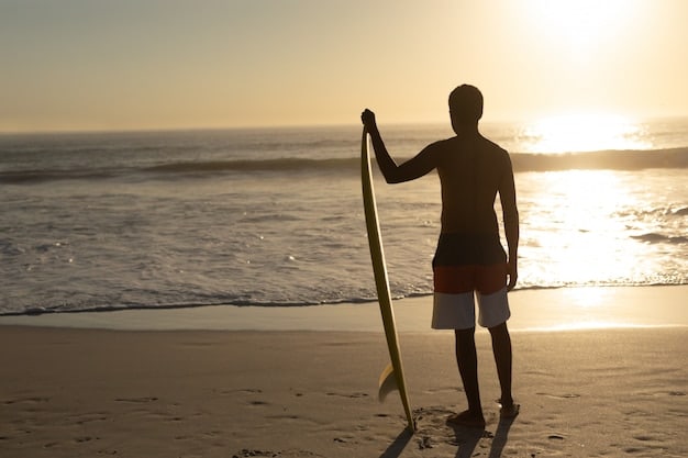 A surfer in a wetsuit cleaning sand off their board with a soft brush after a session, with the ocean and a sunset in the background, implying post-surf care.